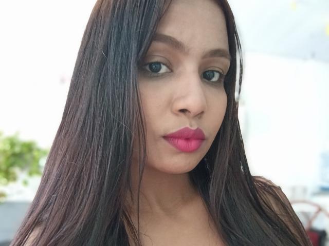 Giuliabe has long, straight brown hair, green eyes, and bright pink lipstick. Her smooth, tan skin and subtle makeup make her face attractive. She's looking down with a pout.