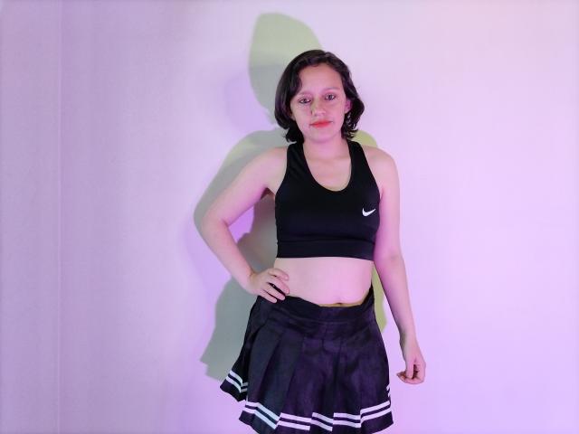 KammiG with medium tits, wearing a black Nike sports bra, black skirt with white stripes, standing against a white wall, hands on hips, short brown hair, neutral expression.