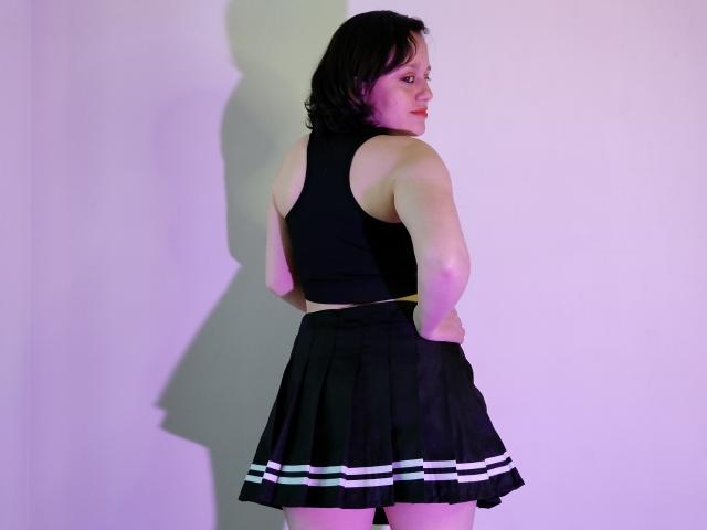 KammiG has short black hair, wearing a black sleeveless top and a black skirt with white stripes, standing with hands on her ass, looking over her shoulder.