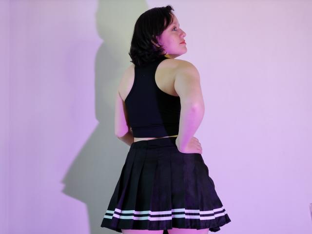 KammiG with black shoulder-length hair, wearing a black sleeveless top and a black skirt with white stripes, standing with hands on hips, looking to the side.