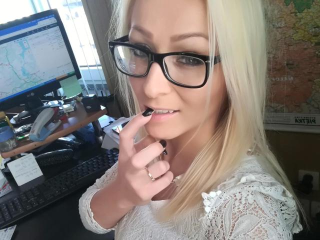 Michaela2015-hot with blonde hair, black glasses, white lace shirt, biting black nail, looking at camera, desk with computer and map in background, smiling slightly.