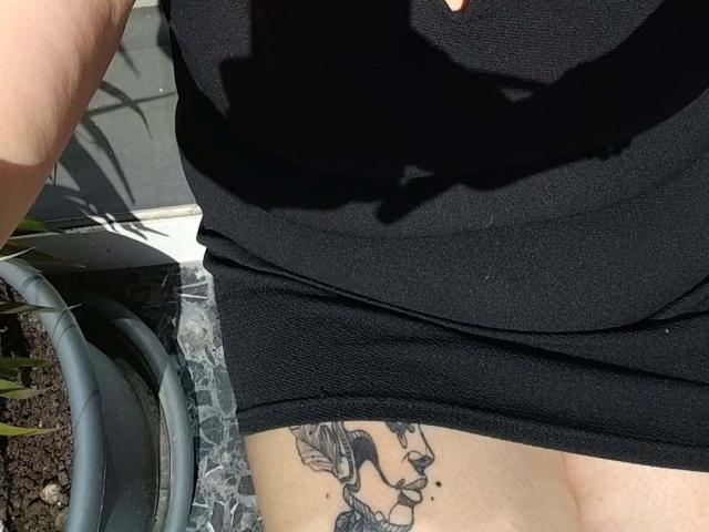 De zwarte jurk van ChaudedeBraise, met een tatoeage van een vrouwen gezicht aan de rechterkant, met een schaduw over haar linkerborst. Zonlicht accentueert haar huid..
