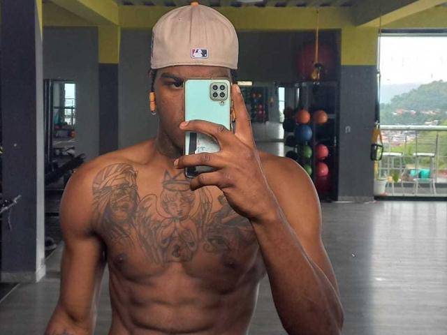 Cataleya e Ethan, senza maglietta, con un petto muscoloso e tatuato, indossano un berretto beige e tengono in mano un telefono che copre il loro viso, si trovano in una palestra con delle palle da ginnastica sullo sfondo..