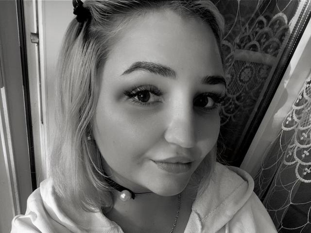 Selfie in bianco e nero di WendyMoira69 con capelli biondi, sopracciglia scure, ciglia, naso piccolo, collana con perla, camicia con cappuccio e un sorriso appena accennato. Il seno di WendyMoira69 non è visibile..
