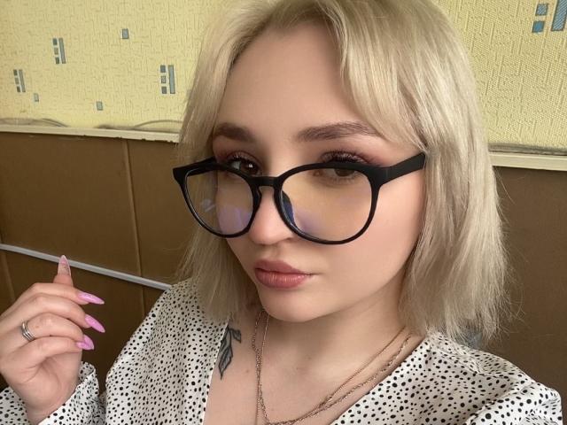 WendyMoira69, com cabelo loiro, óculos de aro preto, unhas cor-de-rosa e uma blusa branca com bolinhas, exibindo uma tatuagem e um anel, com uma aparência fofa e séria..