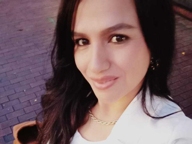CameronViviane has dark brown hair, brown eyes, and is smiling slightly. She's wearing a white shirt with a gold chain necklace, and has a cute, sexy face.