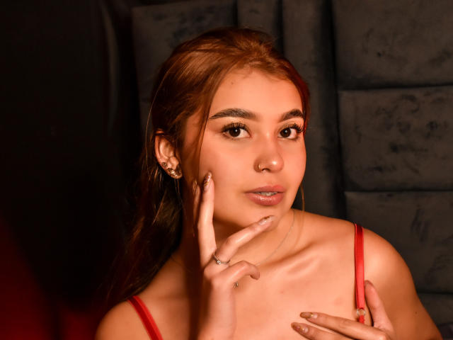 VioletaConnors with light brown hair, red lingerie, and a slight smile, touching her cheek with a ringed finger, brown eyes, and subtle makeup in a warm-lit photograph.