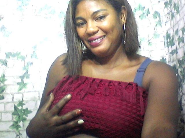 AliceFix with dark skin, brown hair, smiling, wearing a red textured top, holding her tits with dark fingers, pink lipstick, and silver hoop earrings.