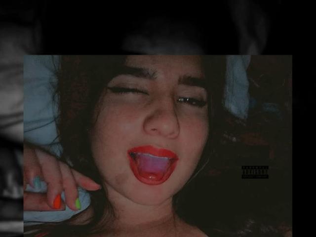 SashaShery has dark hair, her eyes are half-closed, and her red-tipped tongue is out. She has colorful nails and is looking up with a playful expression.