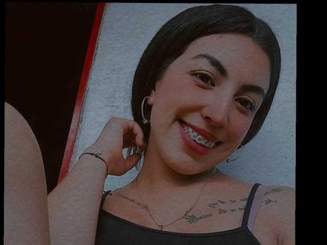 TifanyTobon has dark hair, ear gauges, a smile showing metal teeth, tattoos on her chest, and is wearing a gray tank top with a bracelet on her right hand.