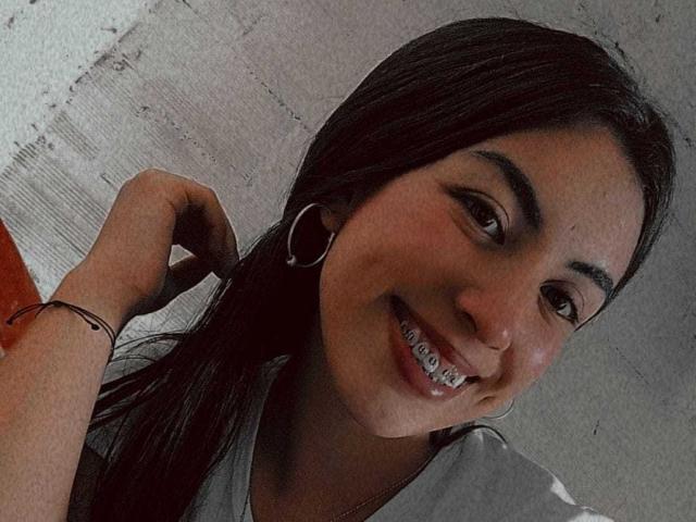TifanyTobon ha i capelli scuri, gli occhi marroni e un grande sorriso con l'apparecchio dentale visibile. Indossa una maglietta grigia, orecchini a cerchio e un braccialetto nero, e si tocca i capelli..