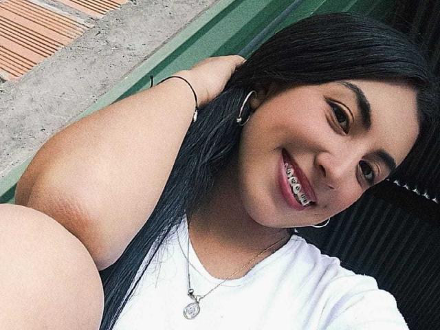 TifanyTobon, con cabello negro, camisa blanca, collar de plata y aros grandes, sonriendo con brackets metálicos, con una mano en el cabello, fondo verde y borde de concreto, mostrando sus glúteos en primer plano..