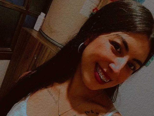 TifanyTobon, con i capelli scuri, sorridente, che mostra i denti, indossa una maglietta bianca, ha un seno piccolo, un tatuaggio sulla spalla sinistra, una collana d'oro e orecchini a cerchio in una stanza poco illuminata..