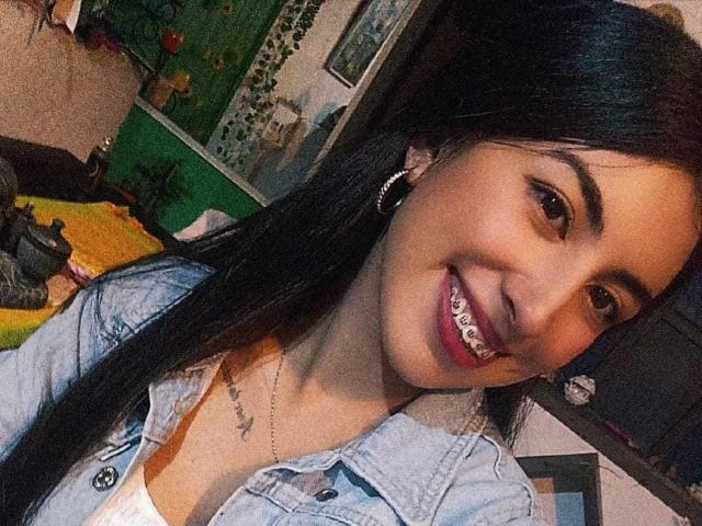 TifanyTobon, com cabelos longos e pretos, sorrindo com aparelho ortodôntico, vestindo uma jaqueta jeans, brincos de argola e um colar, está em pé em um quarto, segurando um selfie..