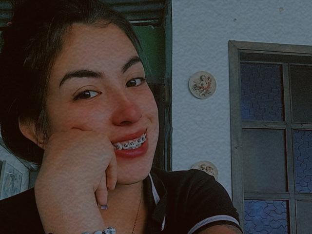 TifanyTobon smiling with her hand on her cheek, showing metal braces on her teeth. She has dark hair, a black shirt, and a light skin tone.