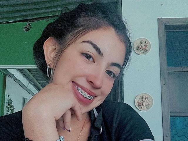 TifanyTobon, com cabelo escuro preso em um coque bagunçado, sorrindo, mostrando aparelho ortodôntico de metal, vestindo uma camisa preta, com a mão no rosto, brincos e colar, em um selfie..
