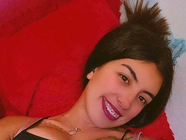 TifanyTobon está acostada sobre un cojín rojo, sonriendo con lápiz labial morado, cabello oscuro recogido en una coleta, y lleva puesto un top negro con tirantes que deja ver sus senos. Lleva puesto un collar..