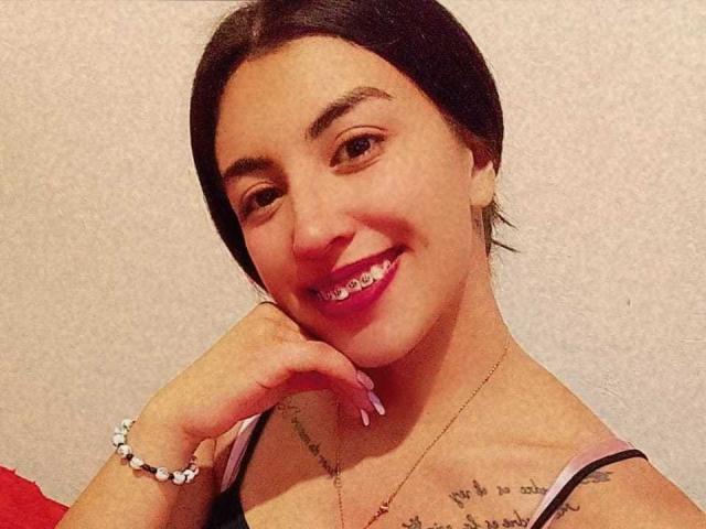 TifanyTobon, con cabello oscuro, sonriendo, lápiz labial rosa, pulsera blanca, collar, tatuajes en el pecho, mano en la barbilla, uñas rosas, camisa roja, piel clara, ojos marrones, cabello ligeramente despeinado..