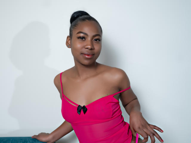 Ariiana Blake, mit dunkler Haut, schwarzen Haaren im Dutt, trägt ein pinkes Kleid mit einem schwarzen Schleifchen, das einen Schulter frei lässt. Sie sitzt, lächelt, zeigt ihre kleinen Brüste und hat rote Nägel..