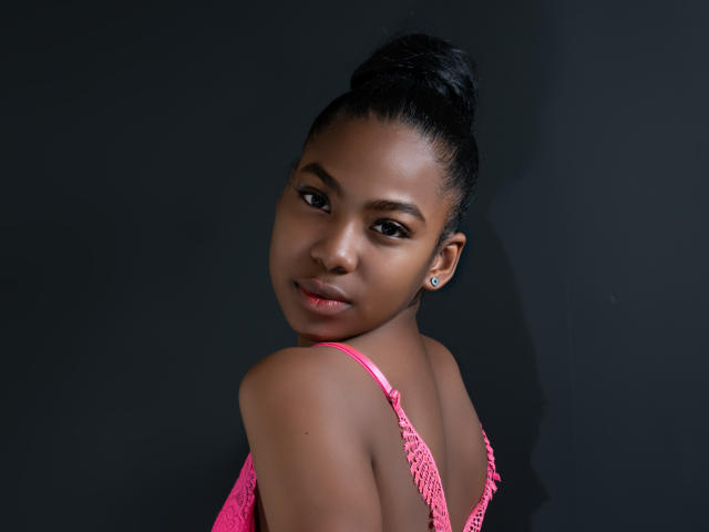 AriianaBlake with dark skin, black hair in a high bun, wearing a pink lace top, looking over her shoulder with a slight smile, showing her ass.