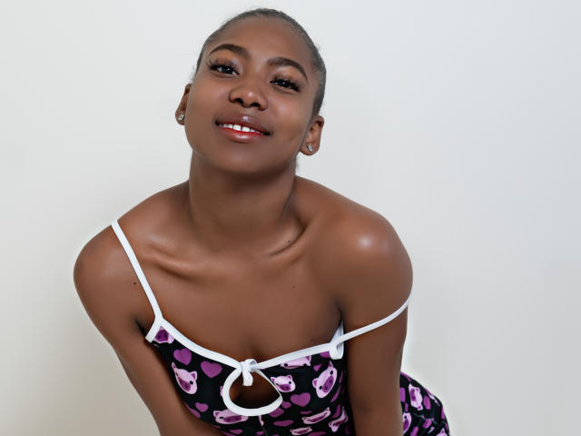 Ariiana Blake, con la pelle scura, sorridente, indossa un vestito nero e viola con un motivo a cuore e spalline bianche, è piegata in avanti, mostrando il suo seno piccolo e un nasino carino..