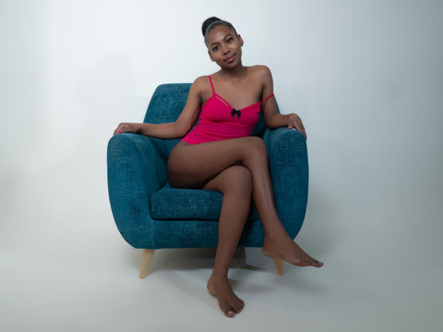 AriianaBlake est assise sur un fauteuil turquoise, vêtue d'un caraco rose avec un nœud noir, qui révèle ses seins et ses fesses, avec les jambes croisées et les pieds nus..