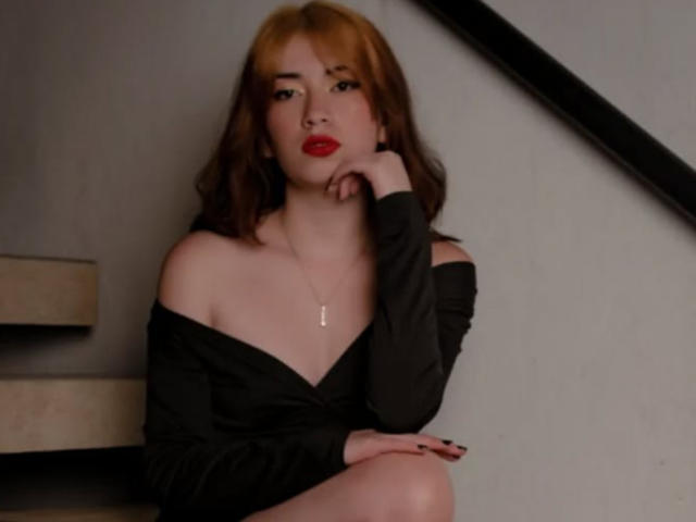 AnyuWu with red lipstick, black off-shoulder top, and brown hair, sitting on stairs, hand on chin, looking at camera, necklace, black nail polish, visible ass, subtle makeup.