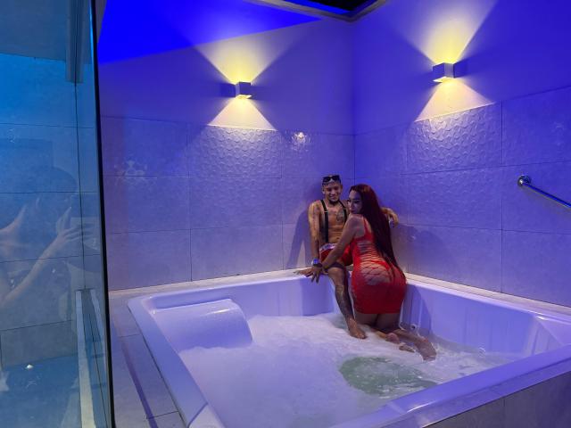 MegaSexyHot man with a muscular build, shirtless, sitting in a white bathtub with bubbles, MegaSexyHot woman in a tight red dress, kneeling, back to camera, water around.