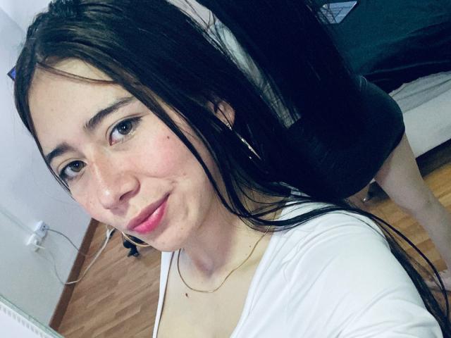 LexyJones mit langen, schwarzen Haaren, einem weißen Oberteil, pinkem Lippenstift, leicht gerötetem Gesicht, kleinen Brüsten, einer goldenen Halskette und goldenen Ohrringen, lächelt in ein Selfie in die Kamera..