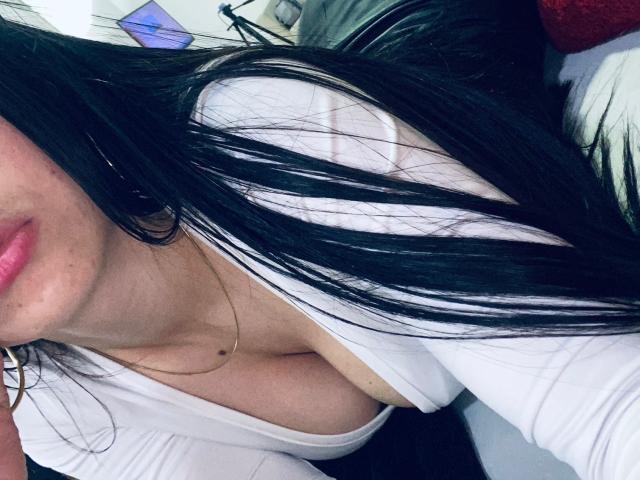 LexyJones, con lunghi capelli neri, labbra rosa e una camicetta bianca che mette in risalto il décolleté, è sdraiata, concentrandosi sulla parte superiore del petto e sul viso, con le labbra rosa leggermente dischiuse, in una luce soffusa..