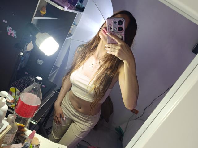LiliiMoore taking a mirror selfie, wearing a white crop top and pants, showing off her tits and flat stomach, with long brown hair, holding a phone with pink case.