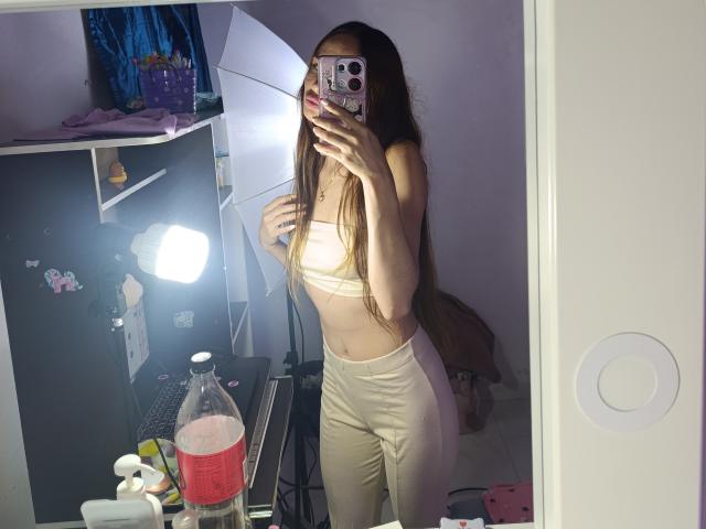 LiliMoore with long brown hair, wearing a white sports bra and high-waisted white pants, taking a mirror selfie, showing her small tits and flat ass, in a dimly lit room.