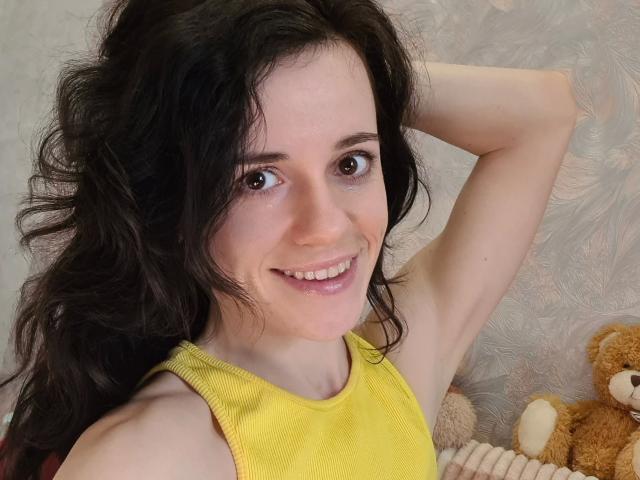 Flower-hot with wavy black hair, brown eyes, smiling, wearing a yellow tank top, arm raised, looking at camera, sitting with a brown teddy bear in the background.
