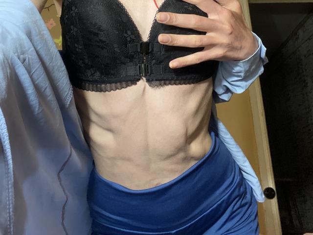 Los abdominales tonificados y musculosos de Kabblasha, con las costillas visibles, lleva un sujetador de encaje negro y una falda azul, sosteniendo el sujetador con una mano, una camisa blanca parcialmente puesta, en una habitación con poca luz..