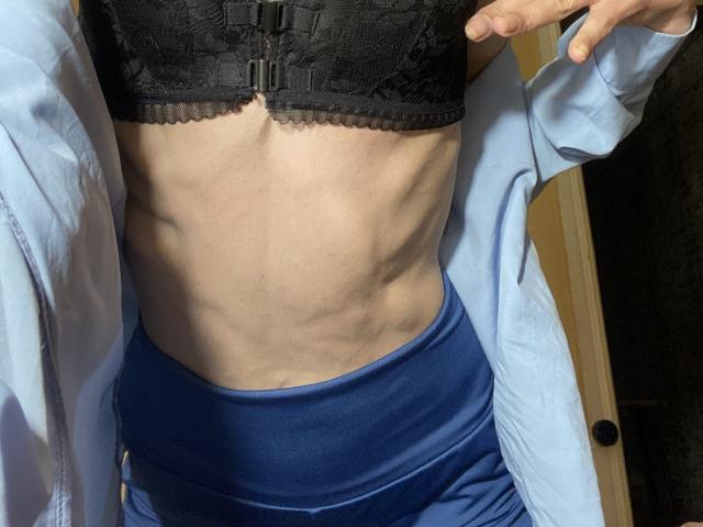 O torso esguio e musculoso de Kabblasha, com costelas visíveis, vestindo um sutiã de renda preto, calças azuis de cintura alta e uma camisa azul clara aberta, mostrando uma mão perto do peito..