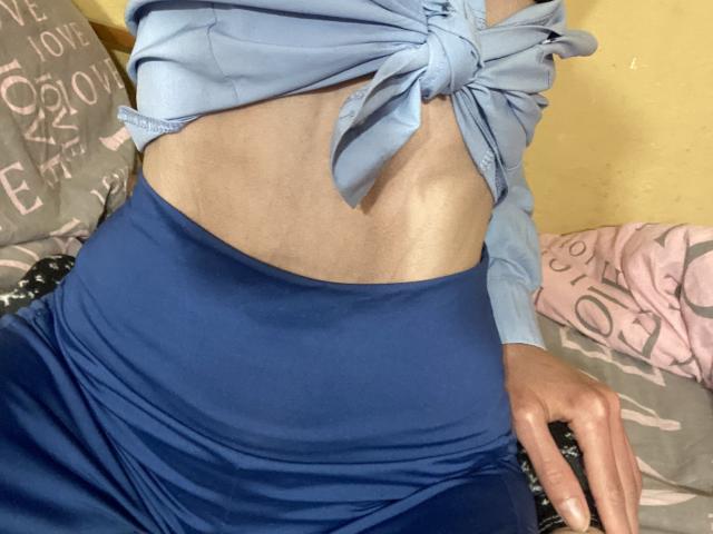 Kabblasha's middelgedeelte, met een blauw topje met een strik dat haar blote buik laat zien, en blauwe broeken met hoge taille, terwijl haar hand op het bed rust..