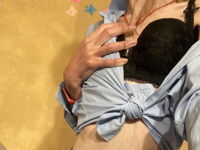 Kabblasha's hand raakt haar zwarte borsten, die bedekt zijn door een beha, terwijl ze een lichtblauwe blouse draagt die dichtgeknoopt is, een rode ketting en een oranje polsband, voor een gele muur..