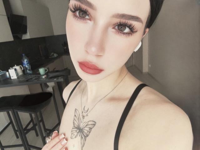 AnayaSweety with pale skin, black hair, and red lipstick, wearing a black spaghetti strap top, showing a butterfly tattoo on her chest, and long eyelashes.