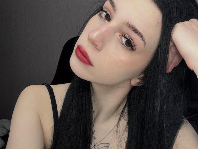 AnayaSweety with pale skin, black hair, red lipstick, and brown eyes, holding her hair up, showing her small tits, and a tattoo on her chest.