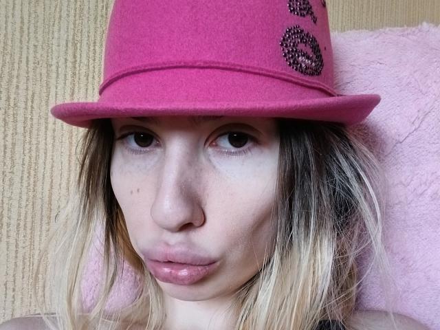 ElzaKomx with pale skin, pouty pink lips, and blonde hair, wearing a pink hat with black bead designs, looking straight at the camera.