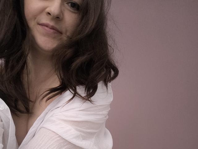 BellLizzie with wavy brown hair, slight smile, wearing a white shirt that reveals cleavage, soft pink background. Her tits are small to medium. She looks cute.