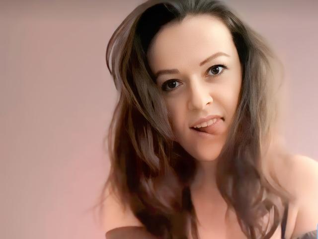 BellLizzie has wavy brown hair, a cute smile, and is sticking her tongue out slightly. She has a fair skin tone and brown eyes. Boobs are partially visible.