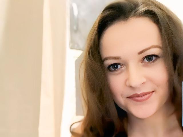 BellLizzie has light brown wavy hair, blue eyes, and a subtle smile with pink lips. Her fair skin is smooth. She's looking directly at the camera.