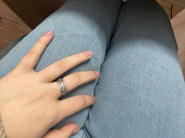 La mano de LaPulia, con un anillo de plata, descansa sobre sus pantalones grises, mostrando sus dedos con uñas rosadas. Sus glúteos están ligeramente separados, revelando la textura de la tela..
