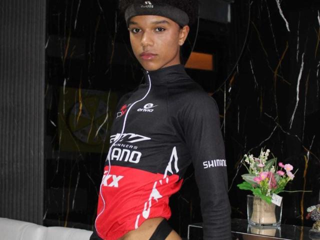 LiamMiranda with short black hair, wearing a black and red triathlon wetsuit, black headband, and black underwear, standing in front of a black marble wall, looking serious.