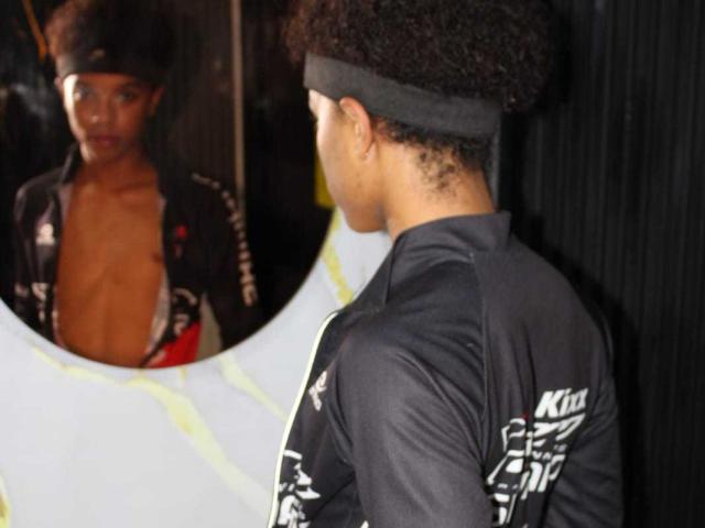 LiamMiranda with curly Afro, black headband, and open black racing suit showing bare chest, reflected in circular mirror, wearing black racing suit with white text.