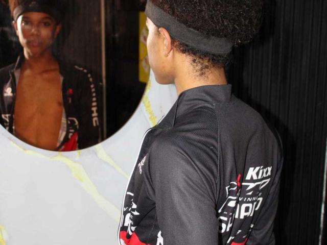 LiamMiranda with curly black hair, wearing a black headband and black sports jacket, facing a mirror reflecting his bare-chested upper body and black jacket.