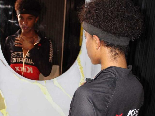 LiamMiranda with curly hair, wearing a black headband and black sports shirt, looks at himself in a mirror. His reflection shows a red and black sports top.