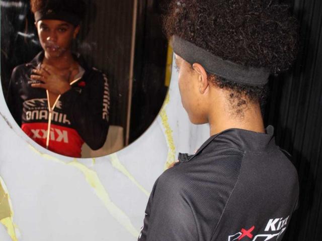 LiamMiranda with curly afro, black headband, and black sports shirt, facing mirror, looking at reflection, holding cigarette, white and yellow taped-up circular mirror.