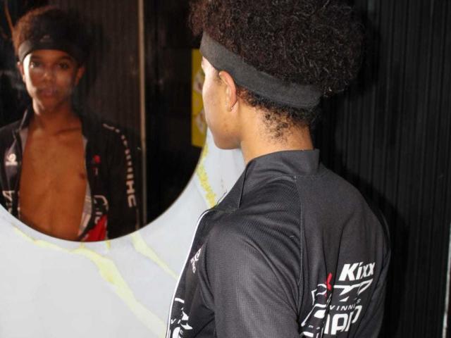 LiamMiranda with curly black hair, wearing a gray headband and black sports shirt, reflects in a mirror showing his bare chest and a black shirt with white patterns.